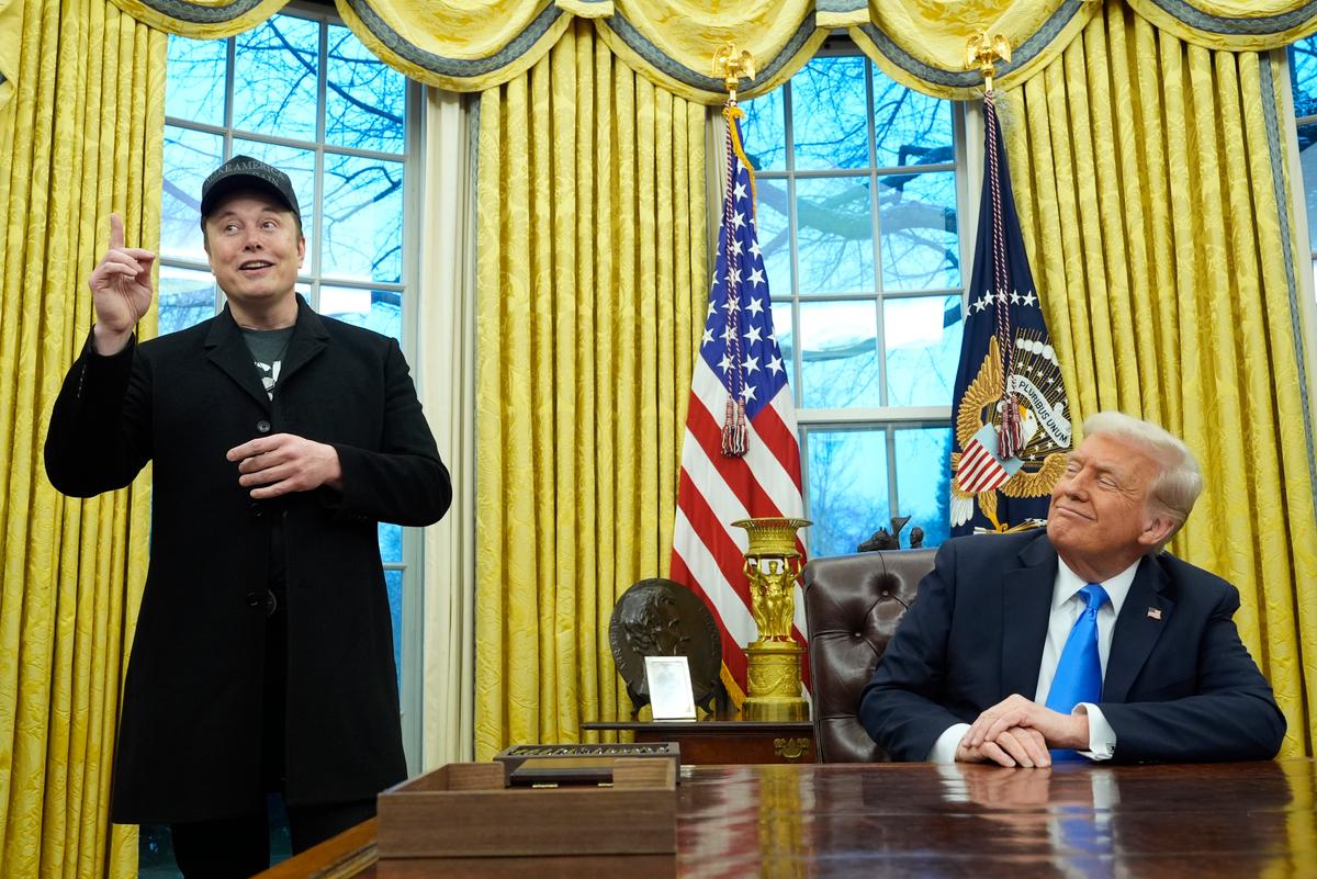 President sitting at his desk, looking at a man in a black coat and ball cap with his finger in the air.